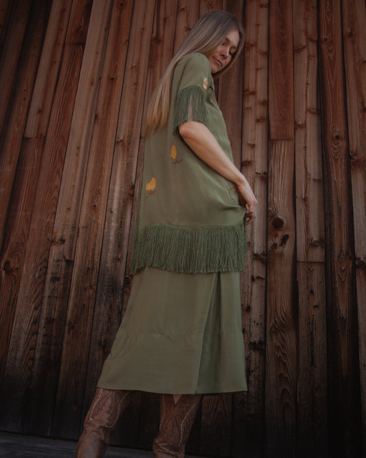 Woman wearing a green dress with fringe against a wooden wall