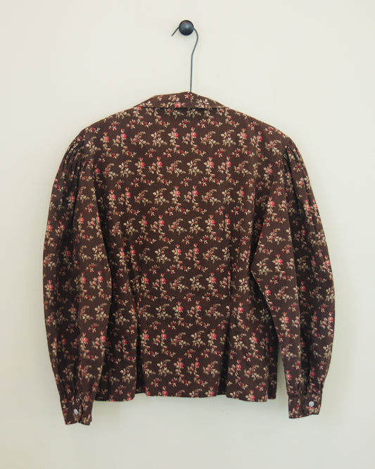 Back of a long-sleeve button-up blouse with a dense small floral print, hanging on a black hanger against a plain off-white wall.
