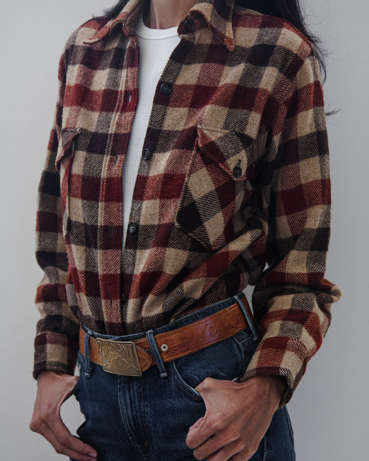 A person wearing a long-sleeve plaid button-up shirt in burgundy, tan, and black over a white t-shirt, paired with blue jeans and a brown leather belt, against a plain light gray background.