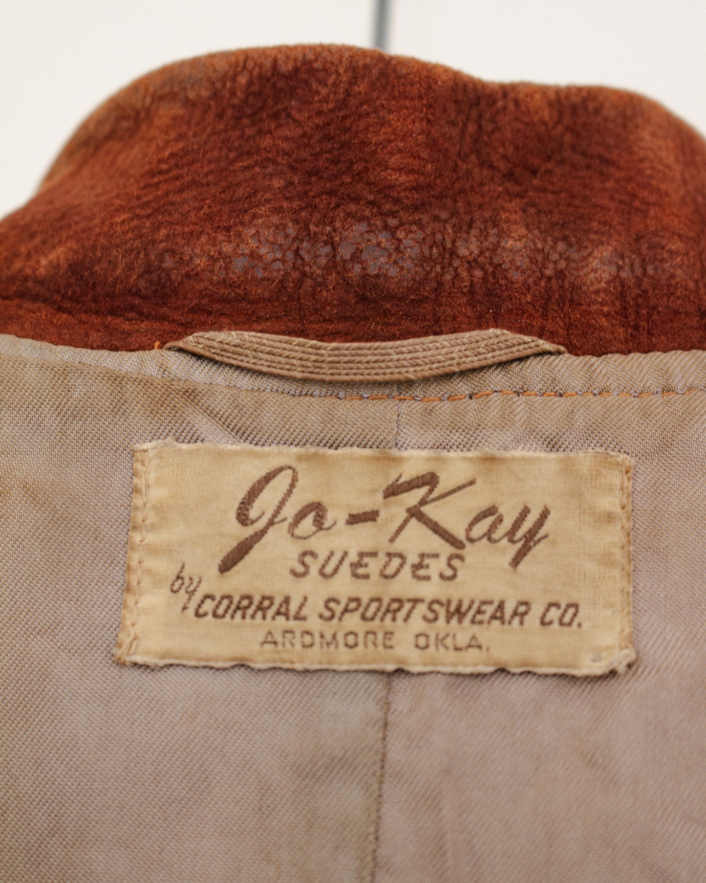 Close-up of the interior neck label of a vintage jacket, showing a beige fabric patch with brown text that reads 'Jo-Kay SUEDES by CORRAL SPORTSWEAR CO. ARDMORE OKLA.