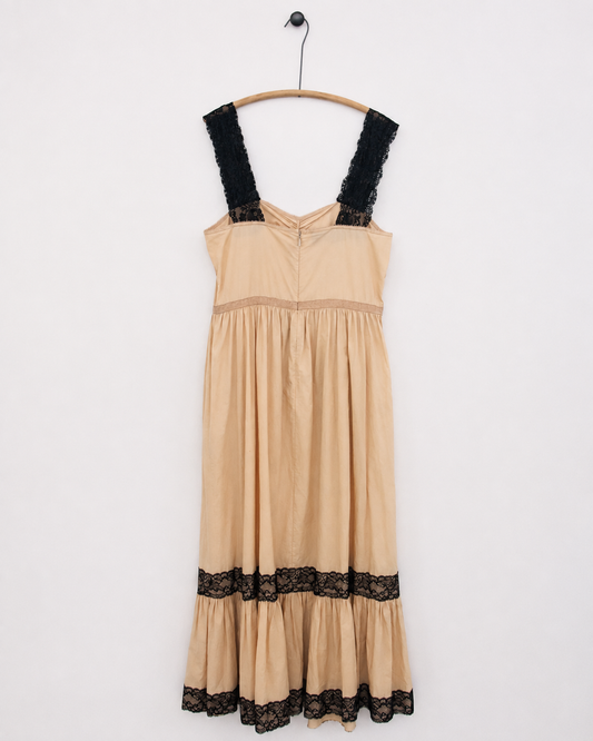 A beige tiered dress with black lace trim on the straps and hem, hanging on a wooden hanger against a plain white wall.