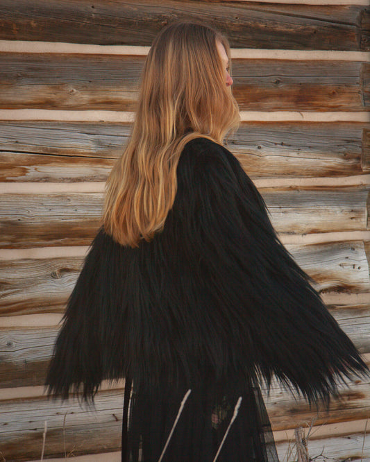 Person wearing a black goat fur jacket standing against a wooden wall.