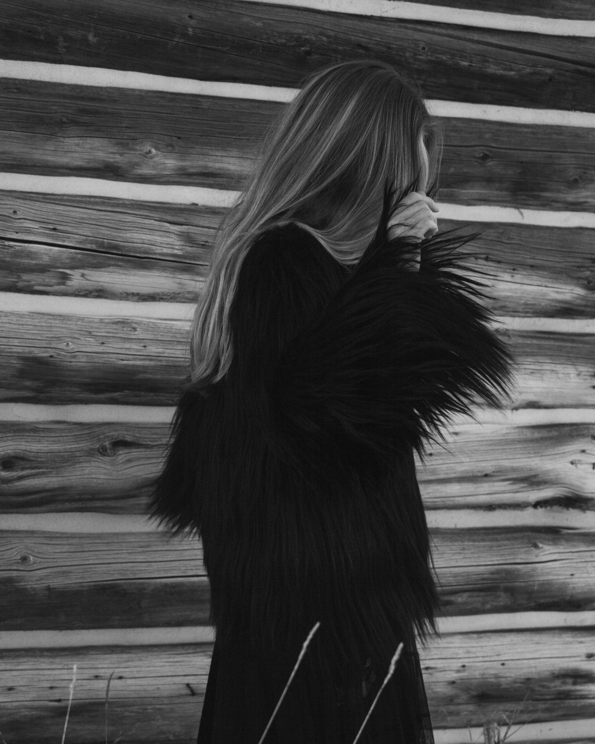 Person wearing a black goat fur jacket standing against a wooden wall.