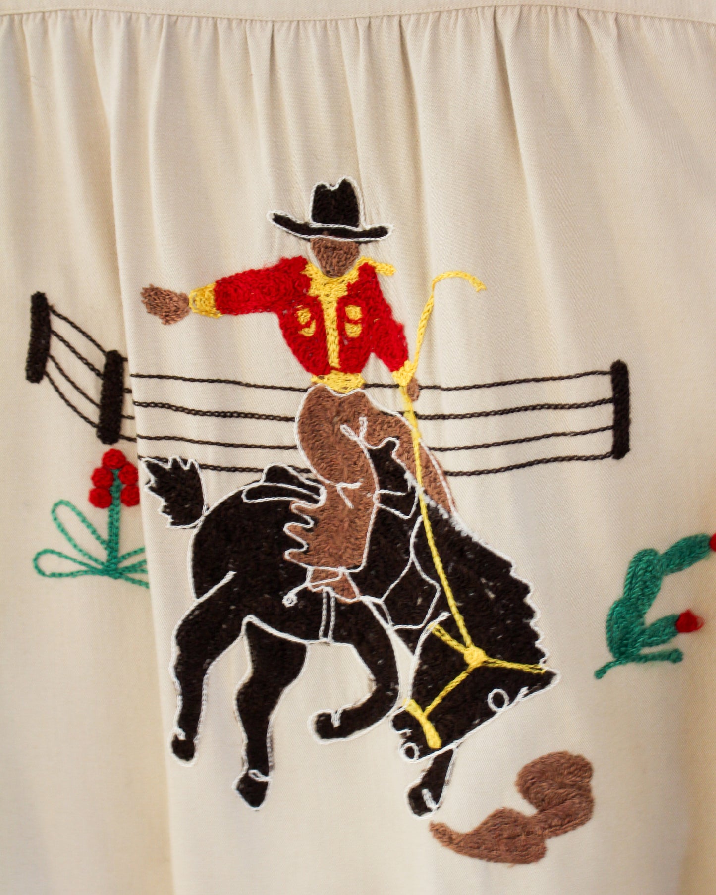 Embroidered design of a cowboy on a horse with a fence and plants on a beige background