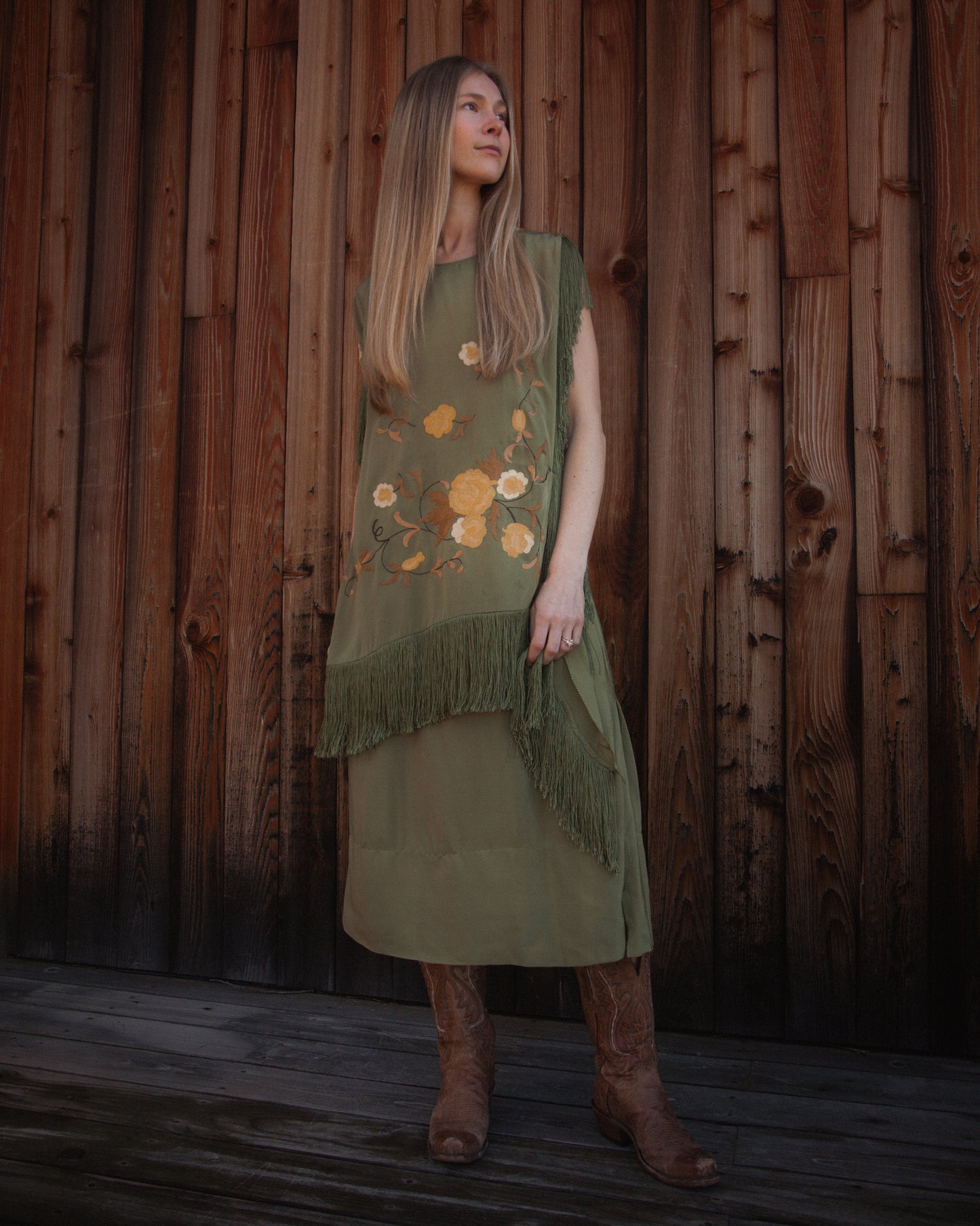 Woman wearing a green dress with floral patterns and fringe against a wooden wall.