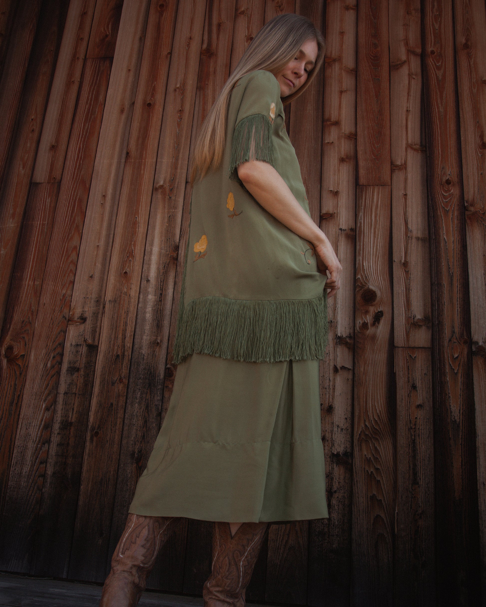 Woman wearing a green dress with fringe against a wooden wall