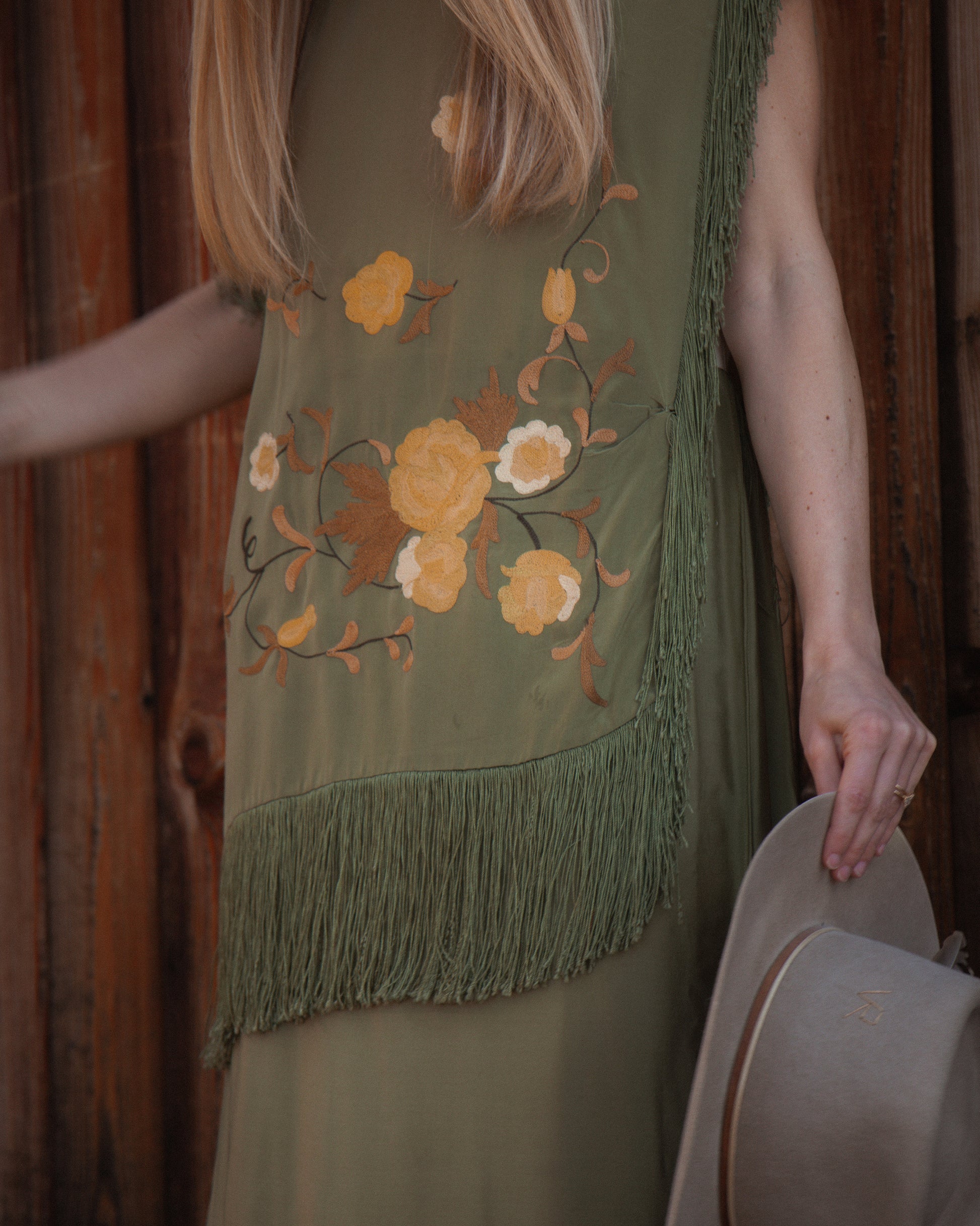Woman wearing a green embroidered dress with floral patterns and fringes, holding a beige hat.