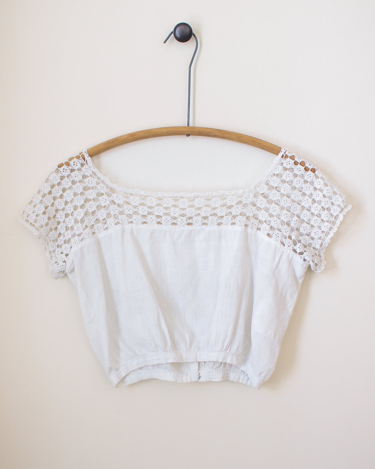 White crop top with lace details on a wooden hanger against a beige wall. View from back