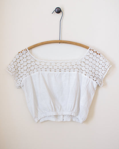 White crop top with lace details on a wooden hanger against a beige wall. View from back