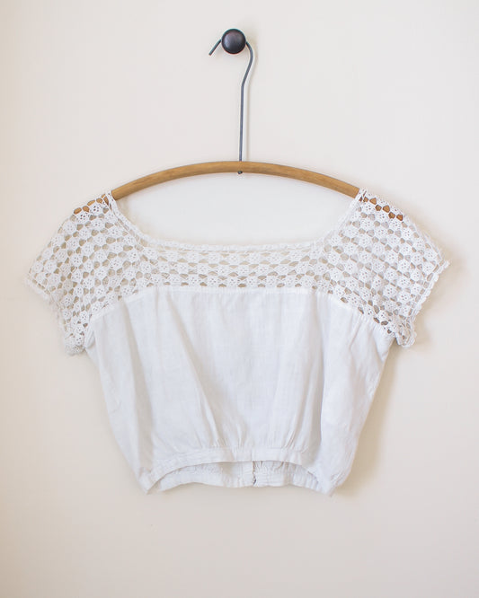 White crop top with lace details on a wooden hanger against a beige wall. View from back