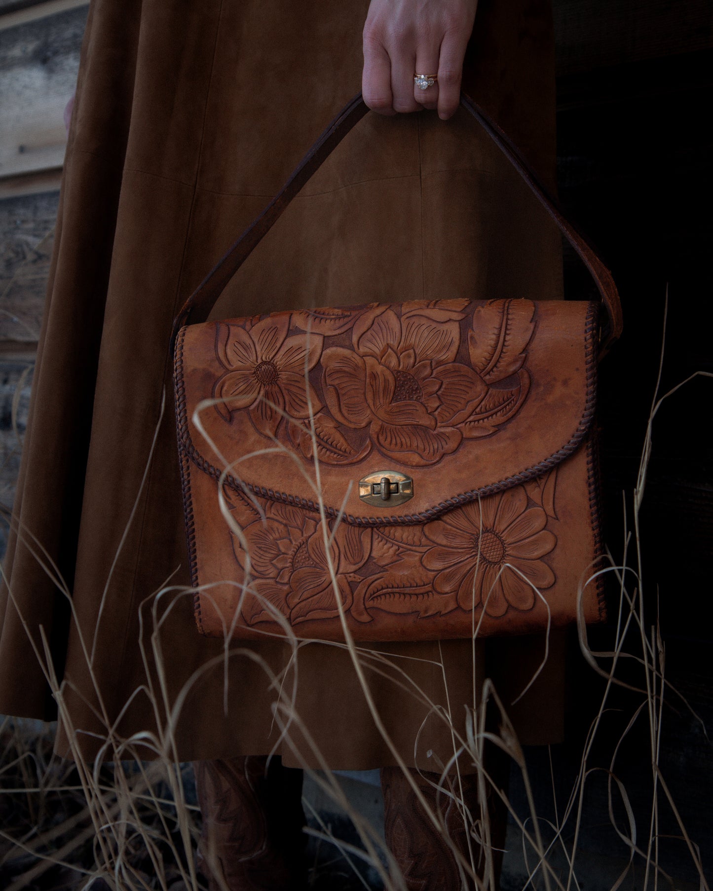 Brown leather handbag with floral patterns held by a person in a coat and boots.