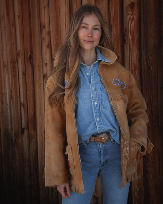 Woman wearing a brown jacket and blue jeans standing against a wooden wall.