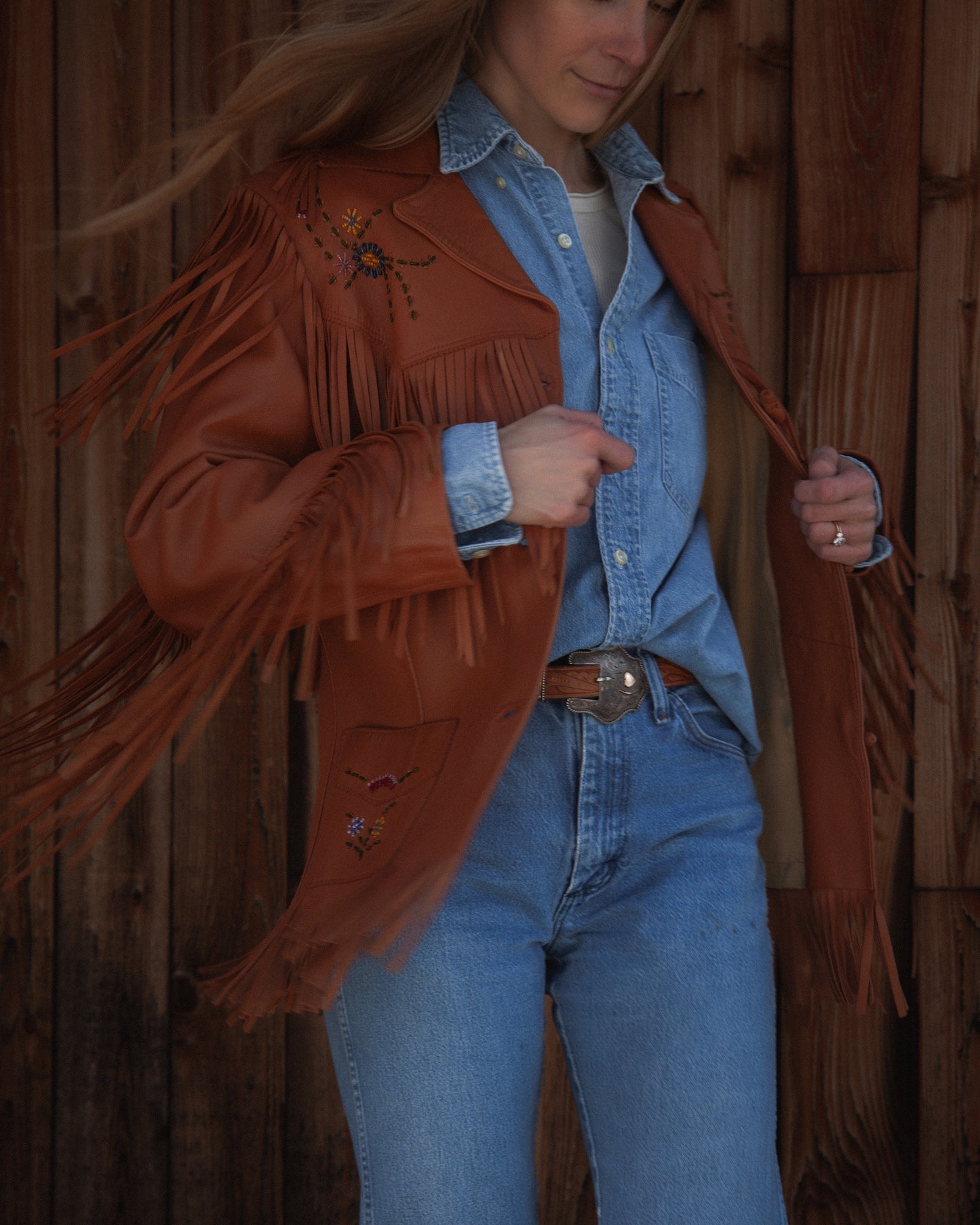 Person wearing a brown fringed jacket with embroidery, blue jeans, and denim shirt against a wooden background.
