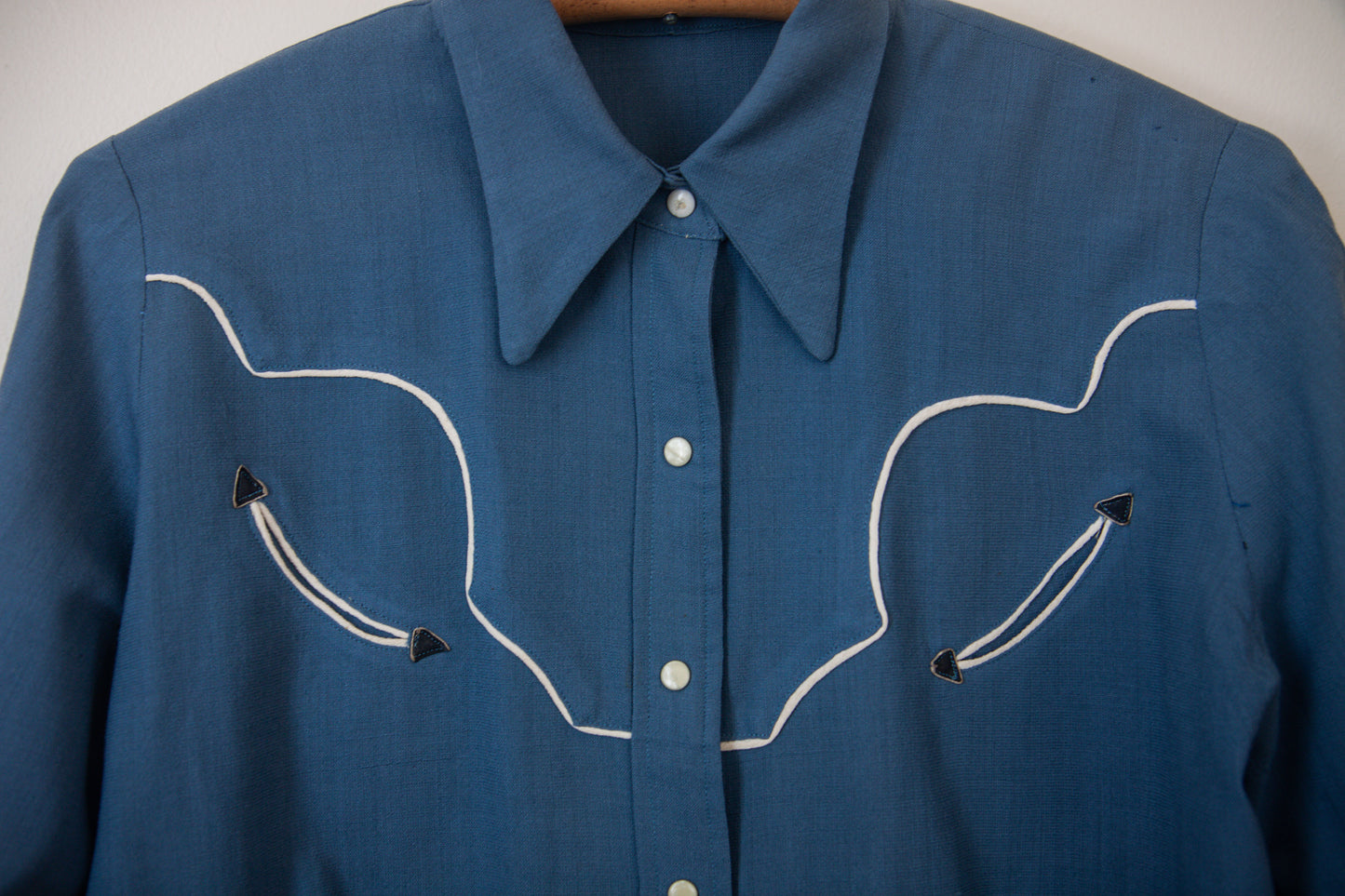 Blue shirt with white embroidery on a hanger against a neutral background