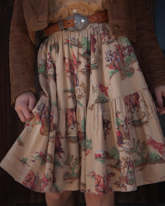 Person wearing a skirt with a colorful pattern of cowboys and horses, paired with a brown jacket.