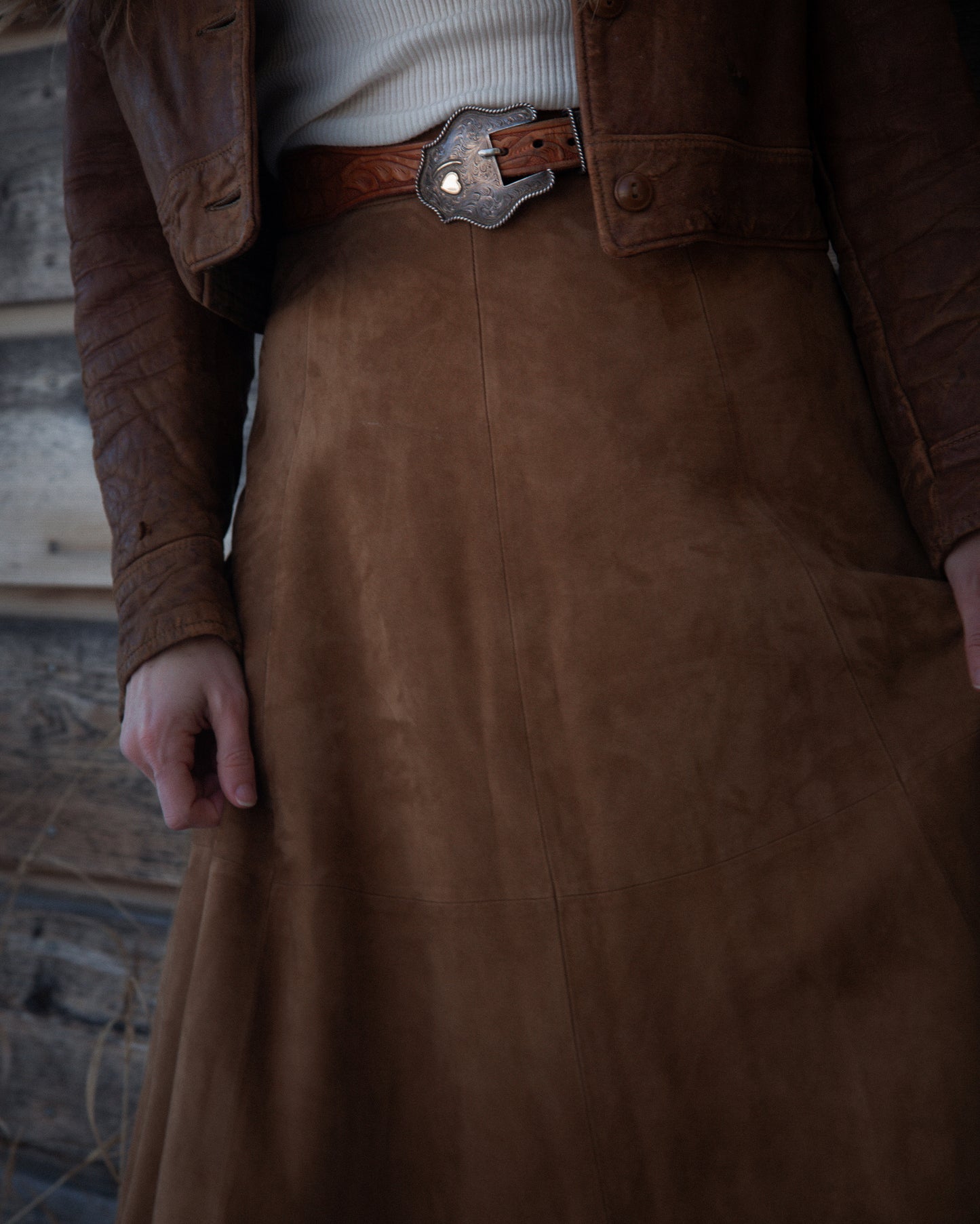 Brown leather jacket and skirt worn by a person against a rustic wooden wall.