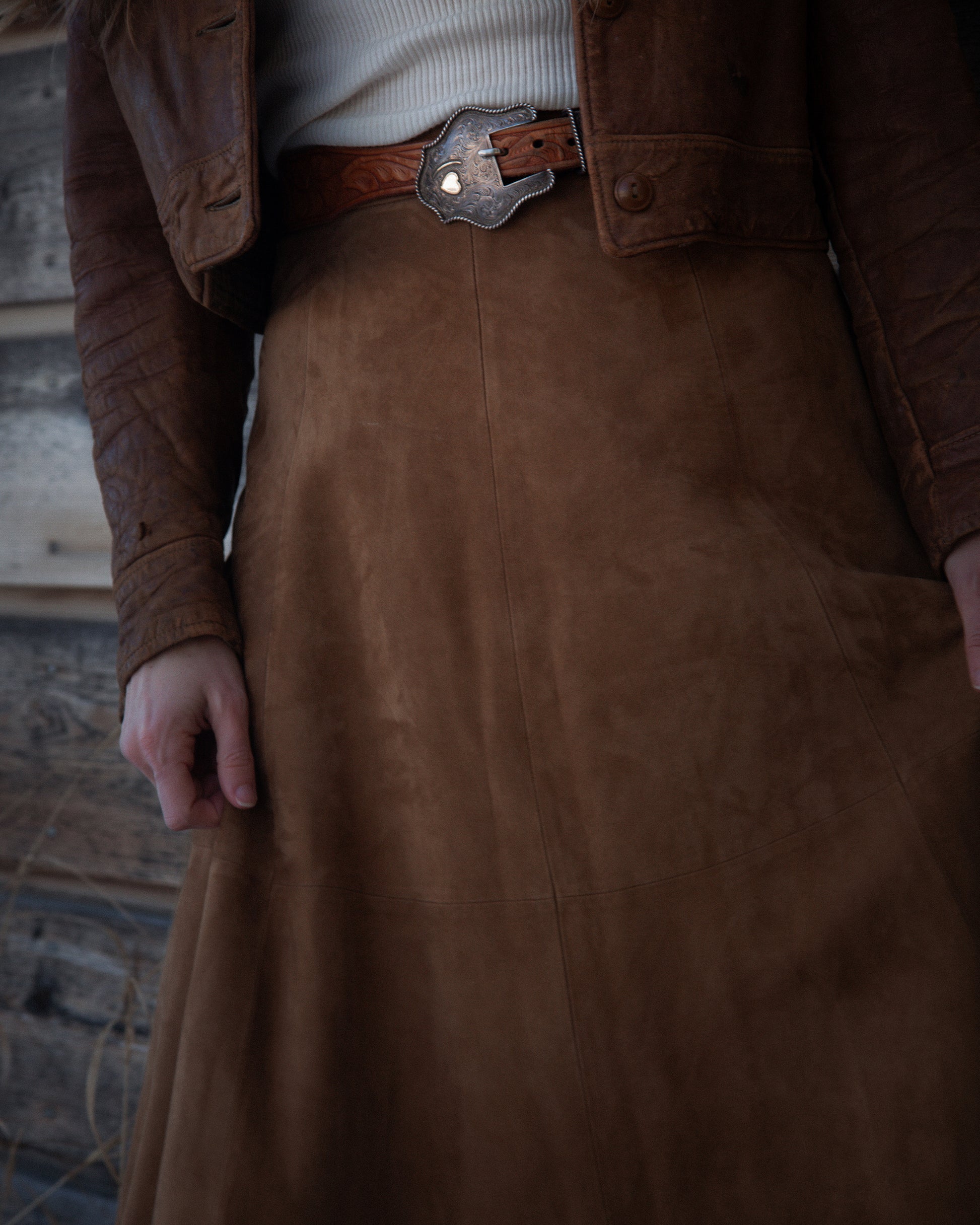 Brown leather jacket and skirt worn by a person against a rustic wooden wall.