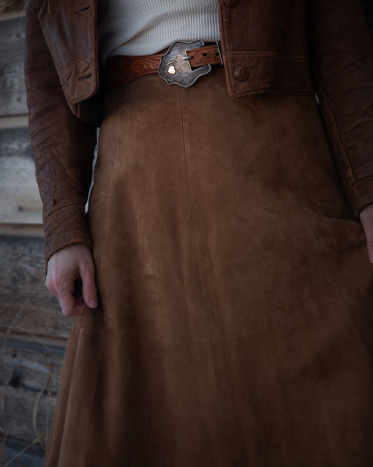 Brown leather jacket and skirt worn by a person against a rustic wooden wall.