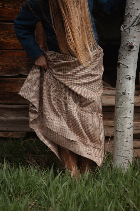 Person wearing brown gingham skirt standing next to a wooden wall and a tree