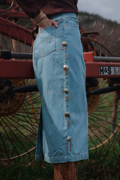 Person wearing a long blue denim skirt with buttons standing next to a large agricultural machine.