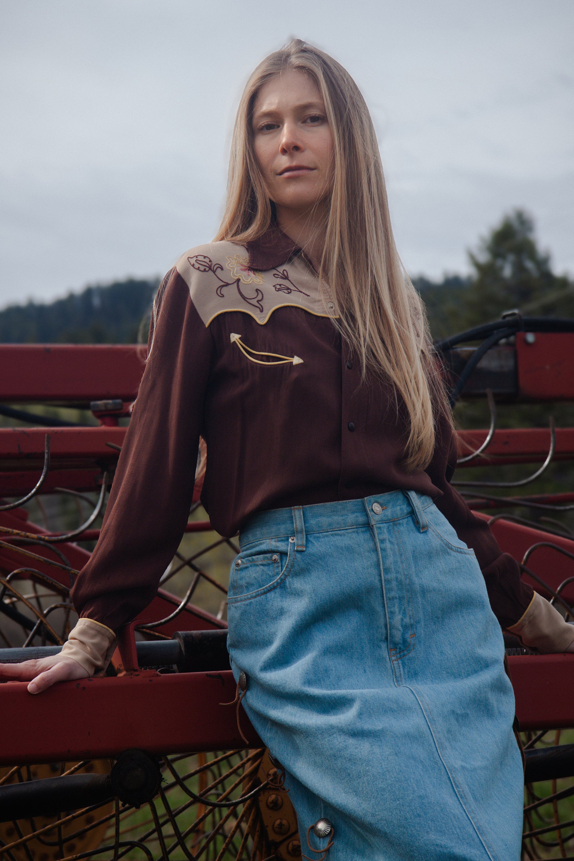Woman wearing a brown blouse with embroidery and blue jeans, standing on a red metal structure.