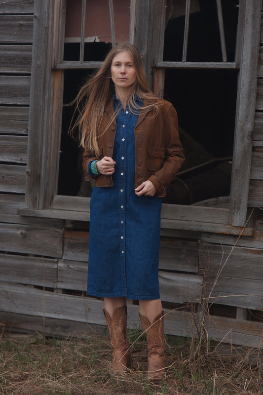 Woman in a blue dress and brown jacket standing in front of an old wooden building.