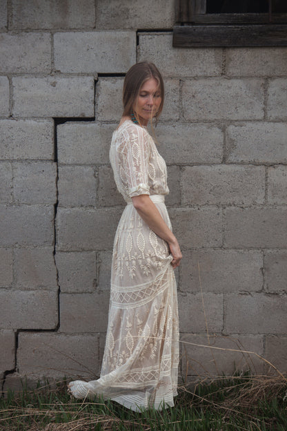 Woman in a white lace dress standing against a gray brick wall.