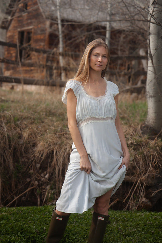 Woman in a white dress standing in a natural setting with a rustic building in the background
