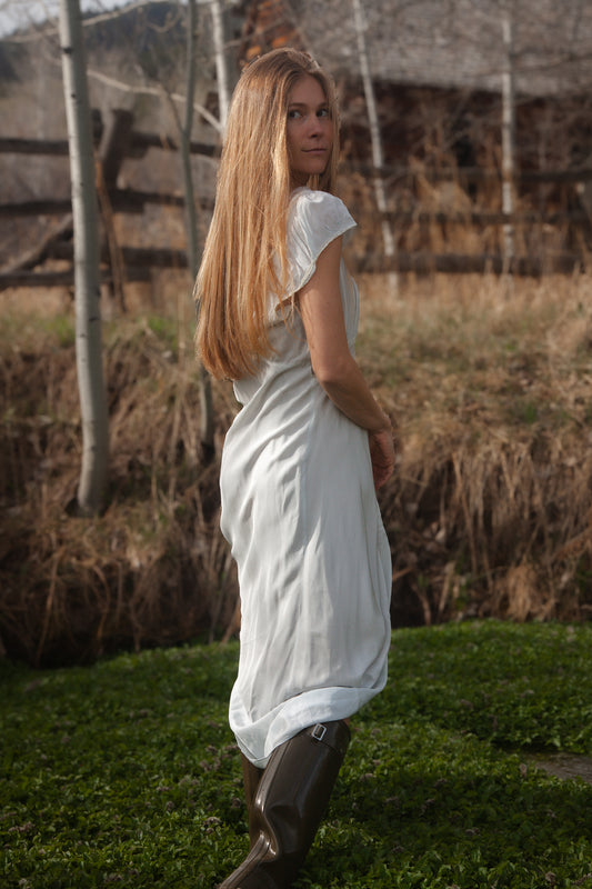 Woman in a white dress standing in a natural setting with trees and grass.