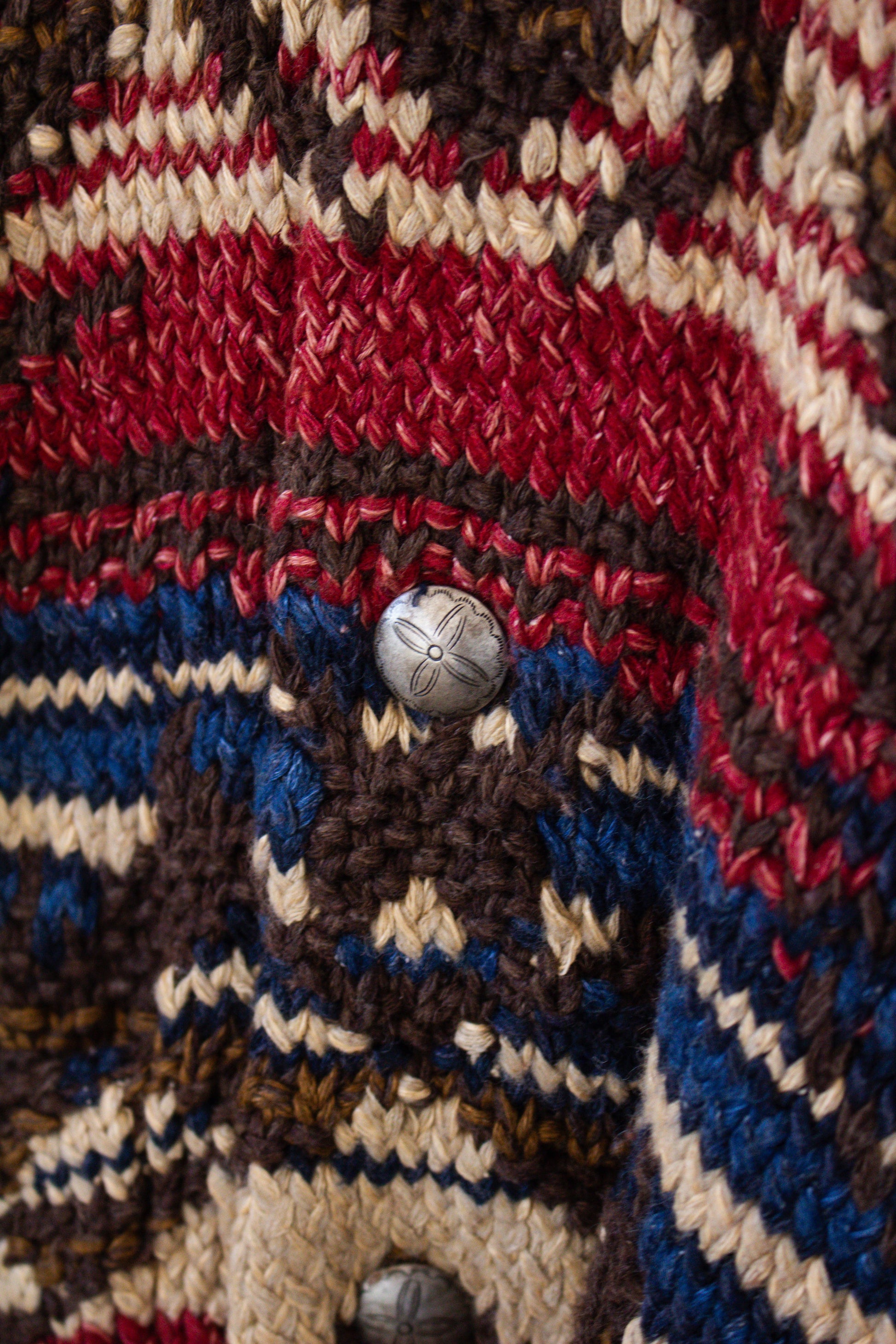 Close-up of a knitted fabric with a button in the center, featuring red, brown, blue, and beige colors.