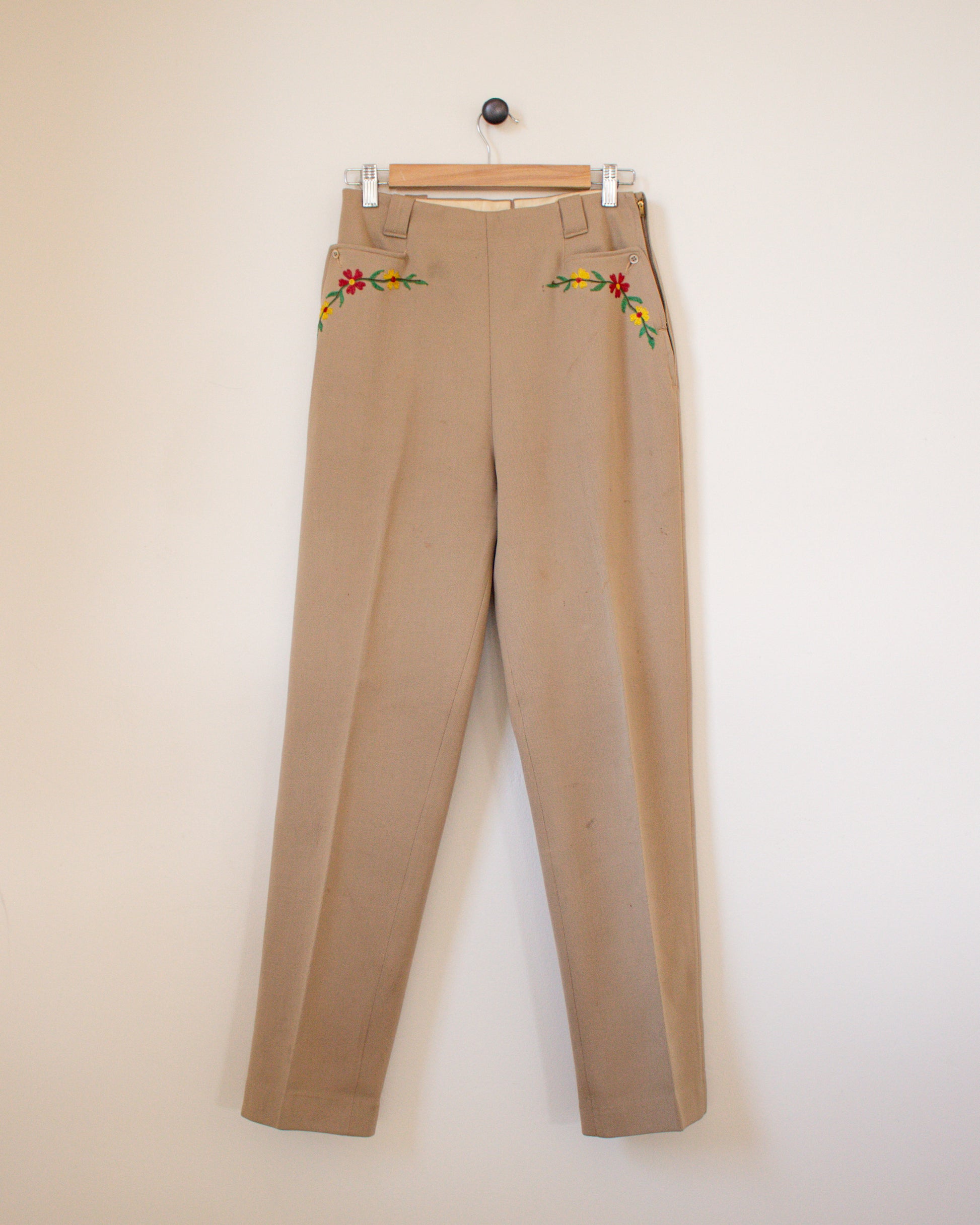 Beige pants with floral embroidery on a hanger against a white background