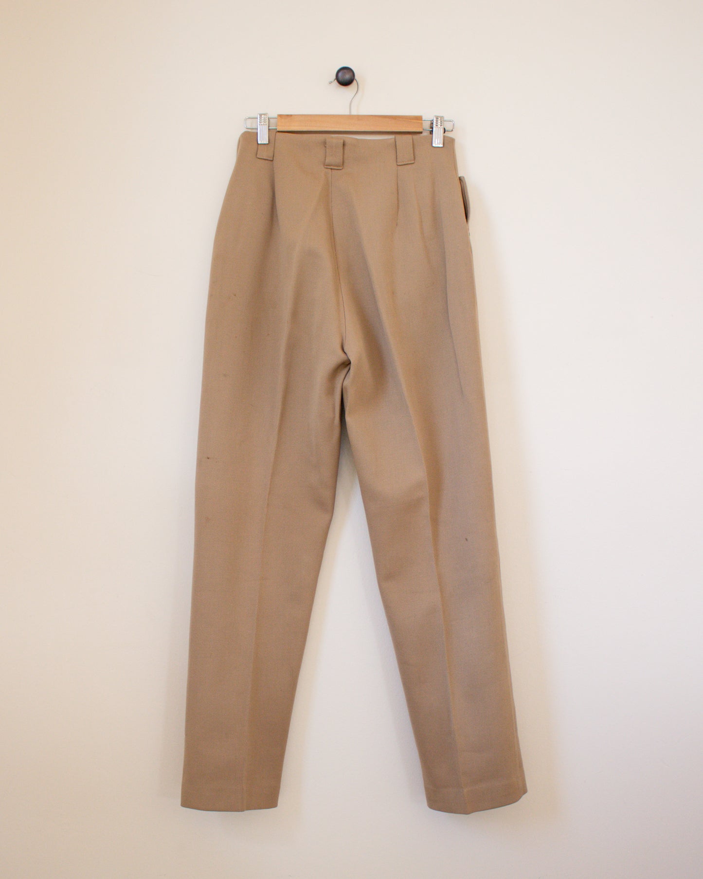 Beige pants hanging on a wooden hanger against a beige wall back view