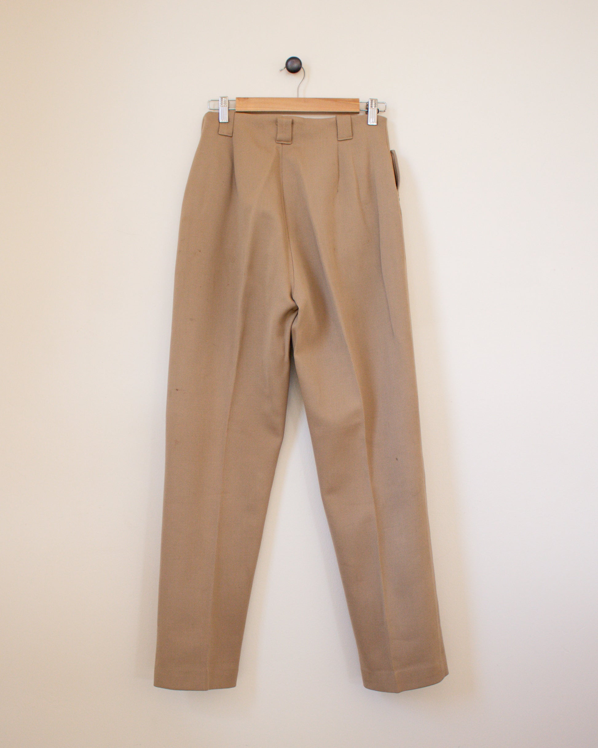 Beige pants hanging on a wooden hanger against a beige wall back view
