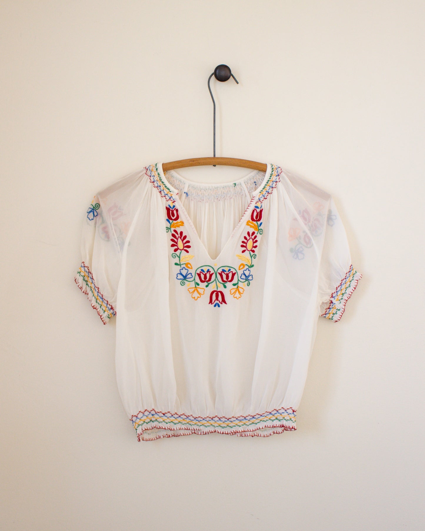 White blouse with colorful embroidery on a hanger against a plain wall