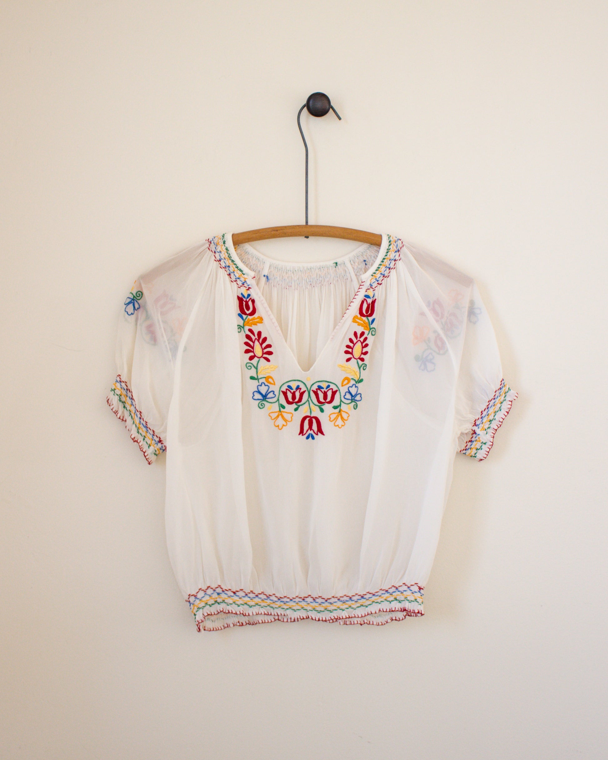White blouse with colorful embroidery on a hanger against a plain wall