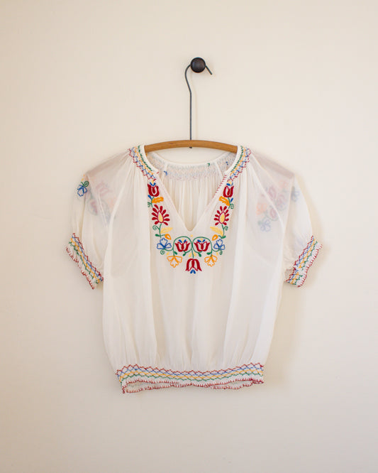 White blouse with colorful embroidery on a hanger against a plain wall