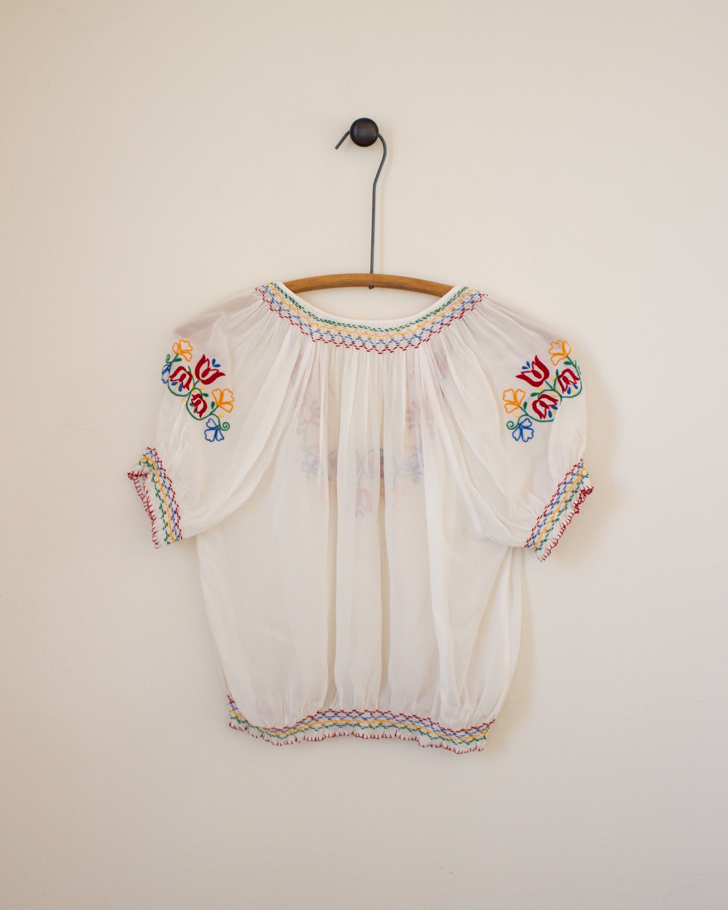 White blouse with colorful embroidery on a wooden hanger against a plain wall.