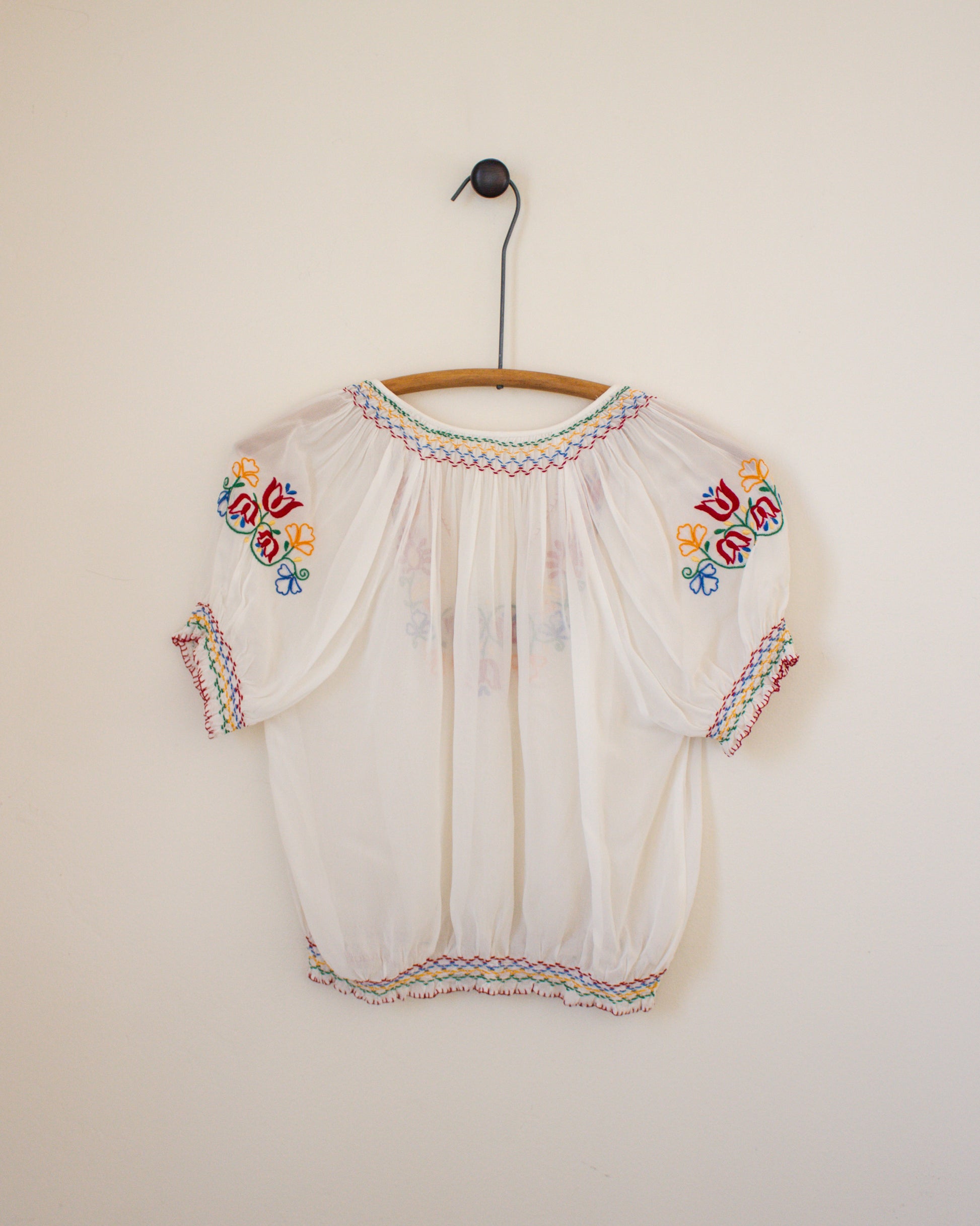 White blouse with colorful embroidery on a wooden hanger against a plain wall.