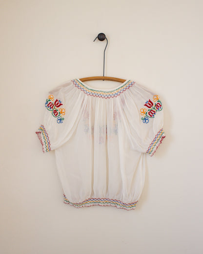 White blouse with colorful embroidery on a wooden hanger against a plain wall.