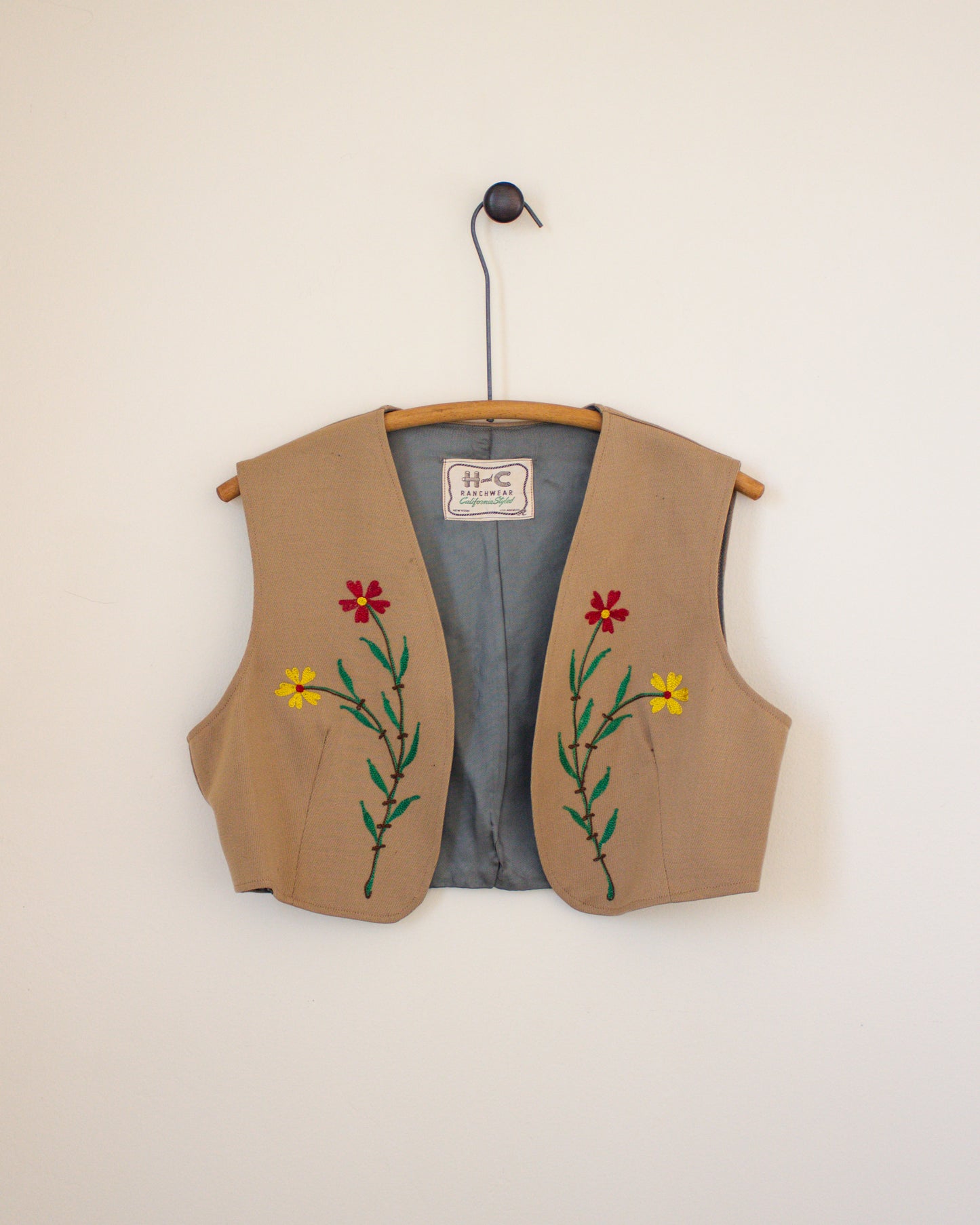 Beige vest with floral embroidery on a hanger against a beige wall