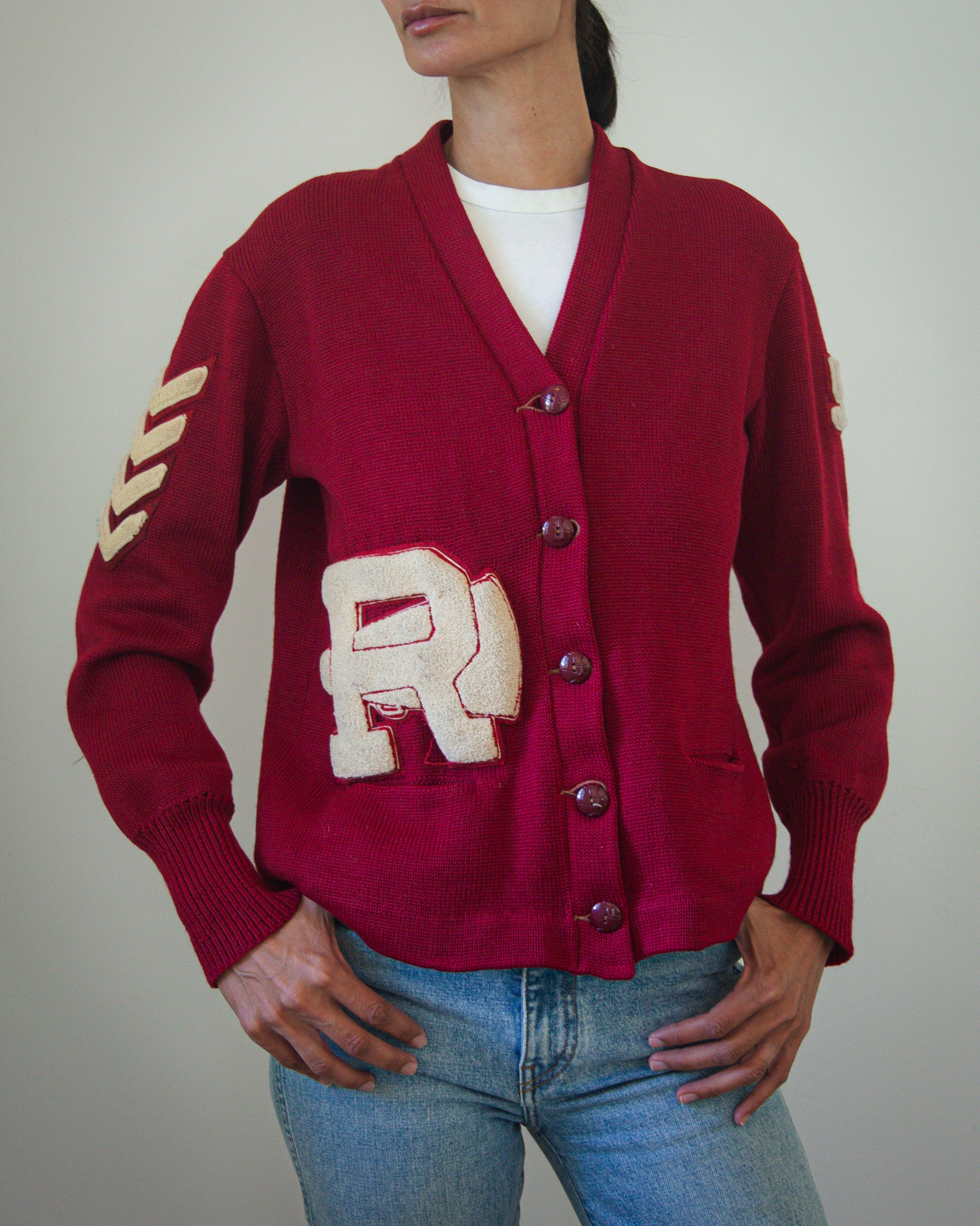 A woman wearing a burgundy knit cardigan with a large cream 'R' patch on the front, matching ribbed cuffs and hem, and cream chevron embroidery on the sleeve, paired with blue jeans.