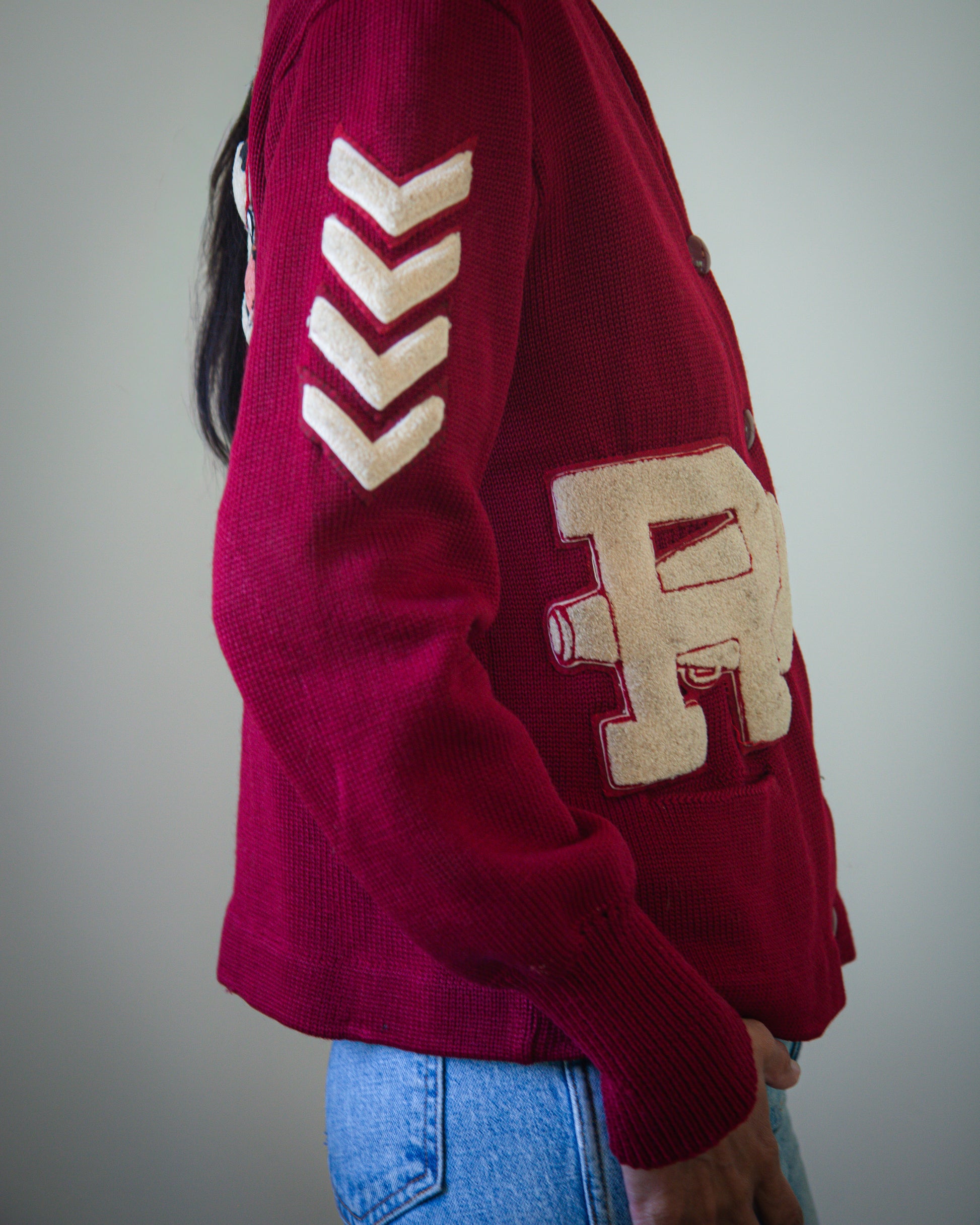 A person wearing a burgundy knit sweater with cream-colored collegiate-style lettering and chevron patches, paired with blue jeans, shown from the side against a plain gray background.