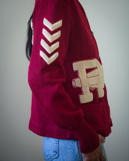 A person wearing a burgundy knit sweater with cream-colored collegiate-style lettering and chevron patches, paired with blue jeans, shown from the side against a plain gray background.