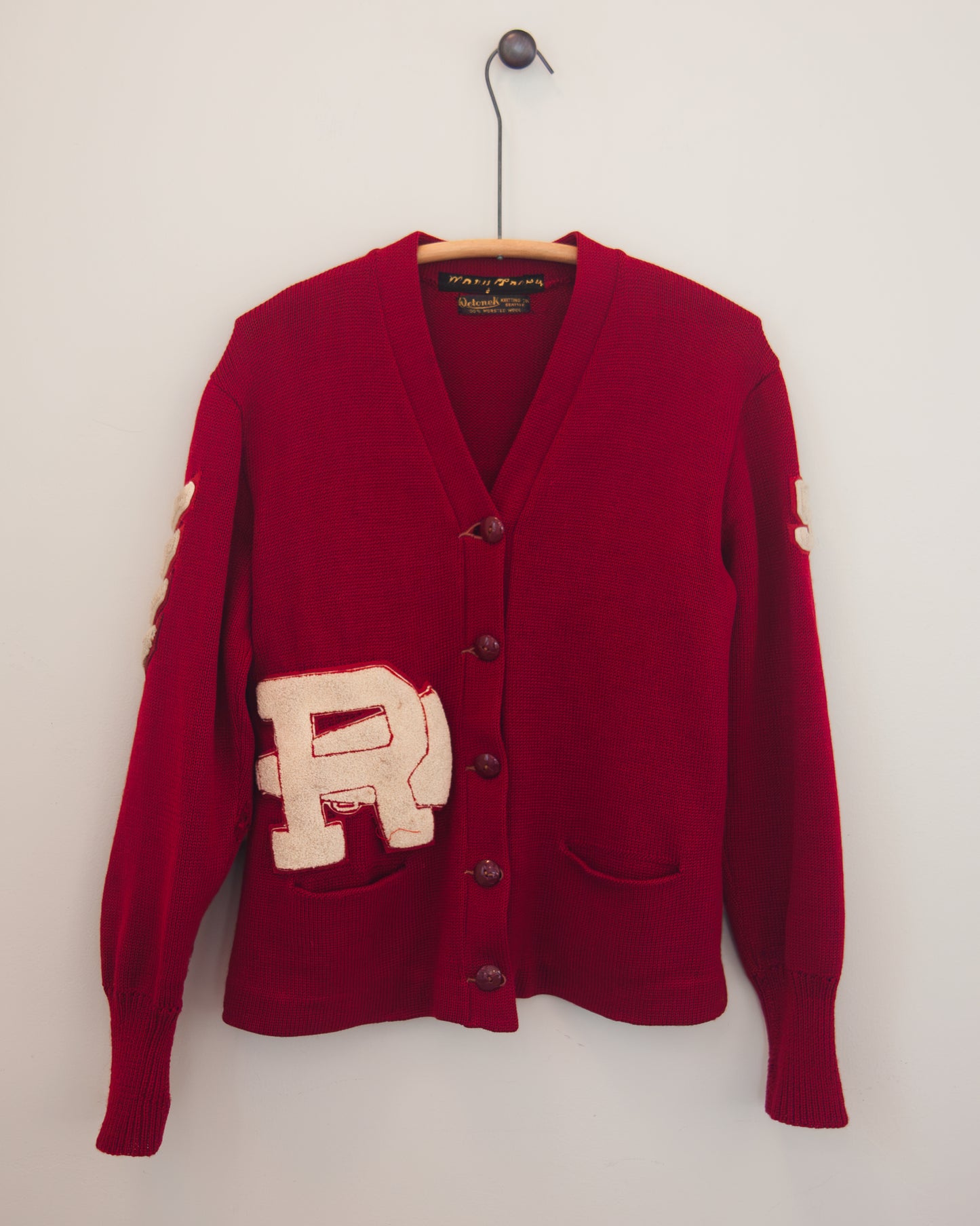 A red knit cardigan sweater hanging on a wooden hanger against a plain white wall. The cardigan features a large beige embroidered letter 'R' patch on the front pocket area, dark brown buttons, and a V-neck.