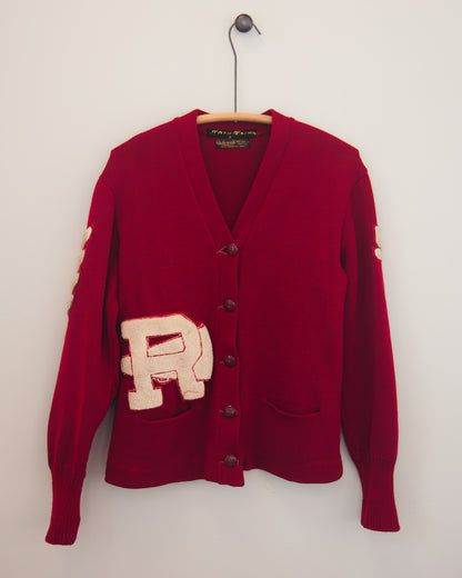 A red knit cardigan sweater hanging on a wooden hanger against a plain white wall. The cardigan features a large beige embroidered letter 'R' patch on the front pocket area, dark brown buttons, and a V-neck.