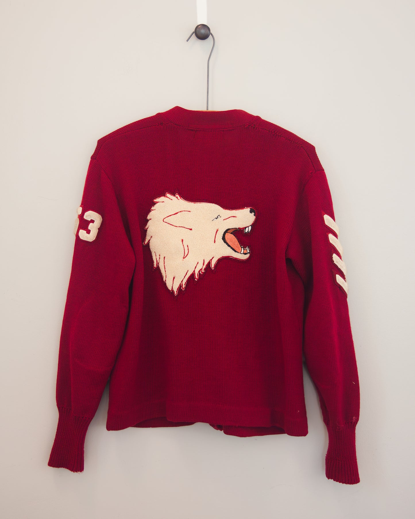 A red knit sweater hanging on a black hanger against a plain light gray wall. The back of the sweater features a large beige embroidered wolf head graphic with its mouth open, and there are additional beige embroidered details on both sleeves.