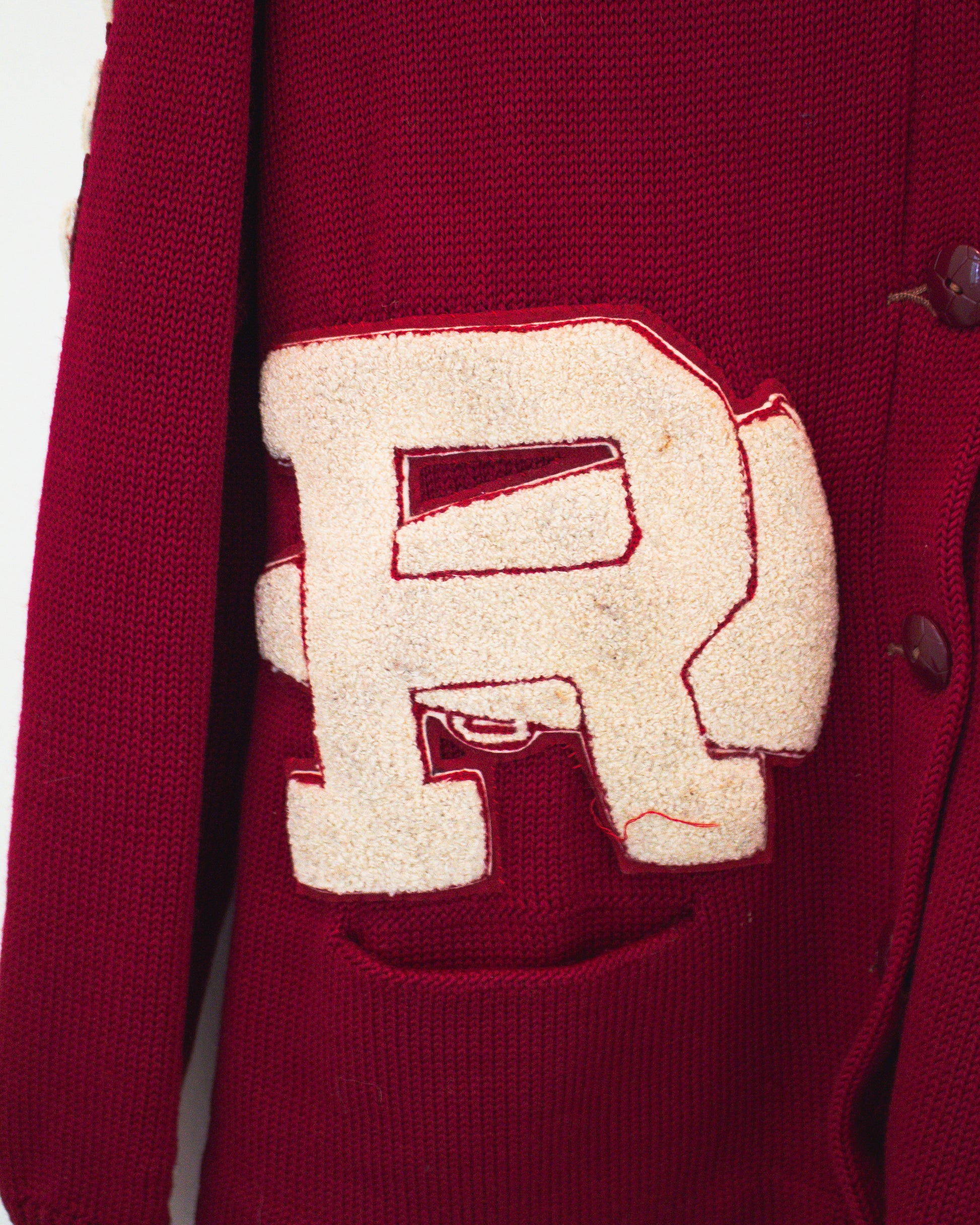 Close-up of a burgundy knit cardigan featuring a large cream-colored embroidered letter 'R' patch on the front above pocket.