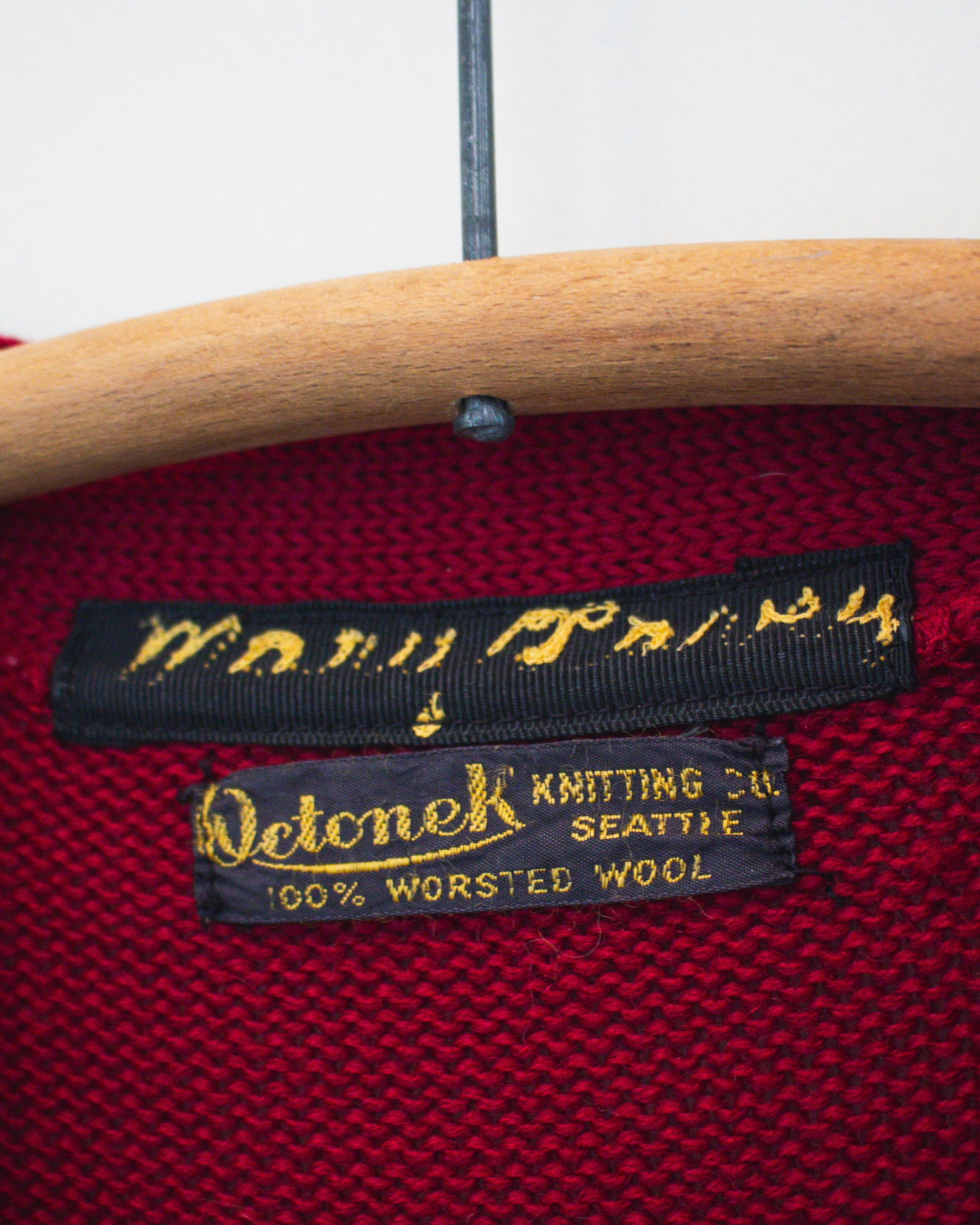 Close-up of a red knit sweater inside label area on a wooden hanger, showing a black sewn-in label that reads Octonek Knitting Co. Seattle, 100% worsted wool.