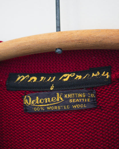 Close-up of a red knit sweater inside label area on a wooden hanger, showing a black sewn-in label that reads Octonek Knitting Co. Seattle, 100% worsted wool.