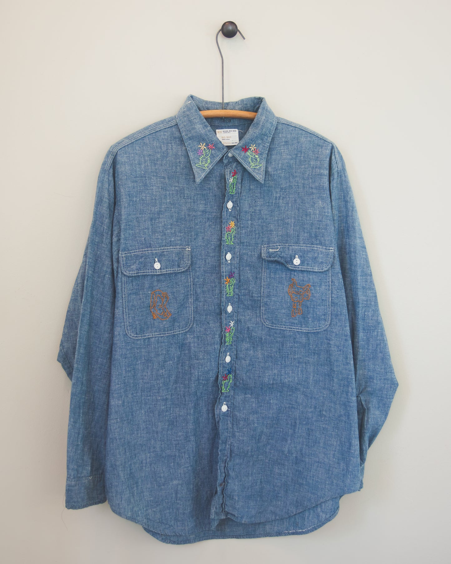 A blue denim long-sleeve button-up shirt with colorful embroidered details on the collar and chest pockets, hanging on a wooden hanger against a plain off-white wall.